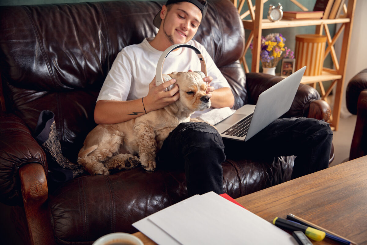 Young handsome man sitting and working at home with his cute dog. Cozy office workplace, remote work, online learning concept.
